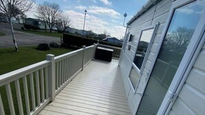 Eine überdachte Terrasse mit weißer Brüstung, einer Schiebetür und Blick auf ein Wohngebiet mit Häusern und blauem Himmel.