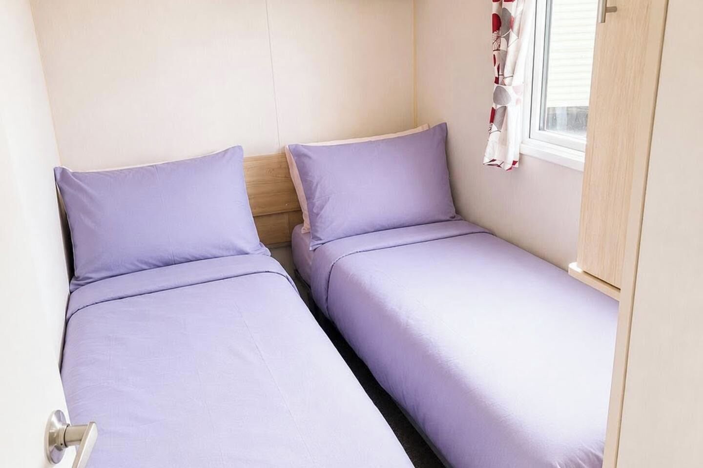 A small room with two single beds, a wooden headboard, a window with blinds, and a door.