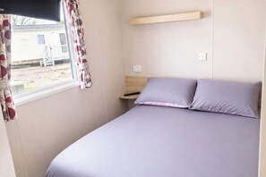 A bedroom with a bed, a nightstand, a window with curtains, and a view of a house outside. - 3 Bed Caravan - Sleeps 8 - Free Parking (Skegness)