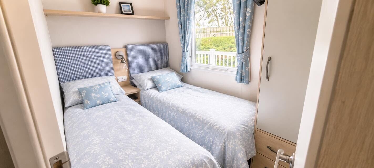 Une chambre à coucher de taille modeste, équipée de deux lits simples, d’une table de chevet avec une lampe et d’une fenêtre avec des rideaux.