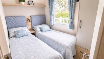 Une chambre à coucher de taille modeste, équipée de deux lits simples, d’une table de chevet avec une lampe et d’une fenêtre avec des rideaux.