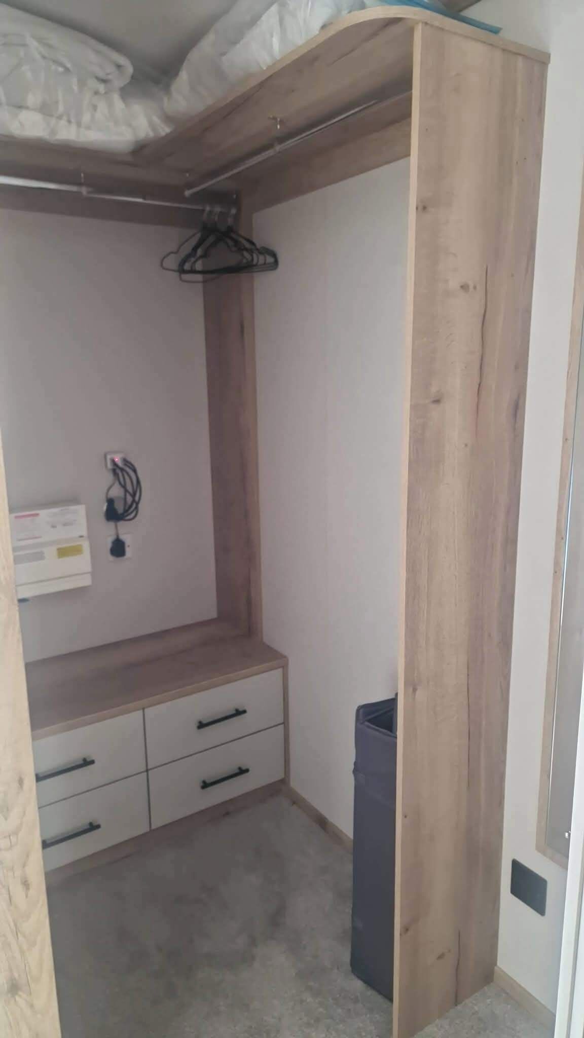 Une pièce comprenant une armoire en bois, un petit bureau avec des tiroirs, un portant à vêtements et une poubelle.