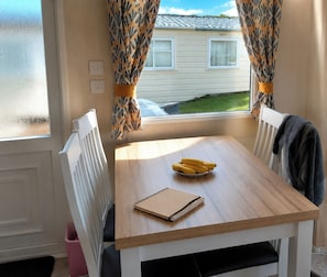 A dining table with a notebook, a bowl of bananas, and a window with curtains. - 3-bed Caravan - Sleeps 8 - Pet Friendly - Parking (Skegness)