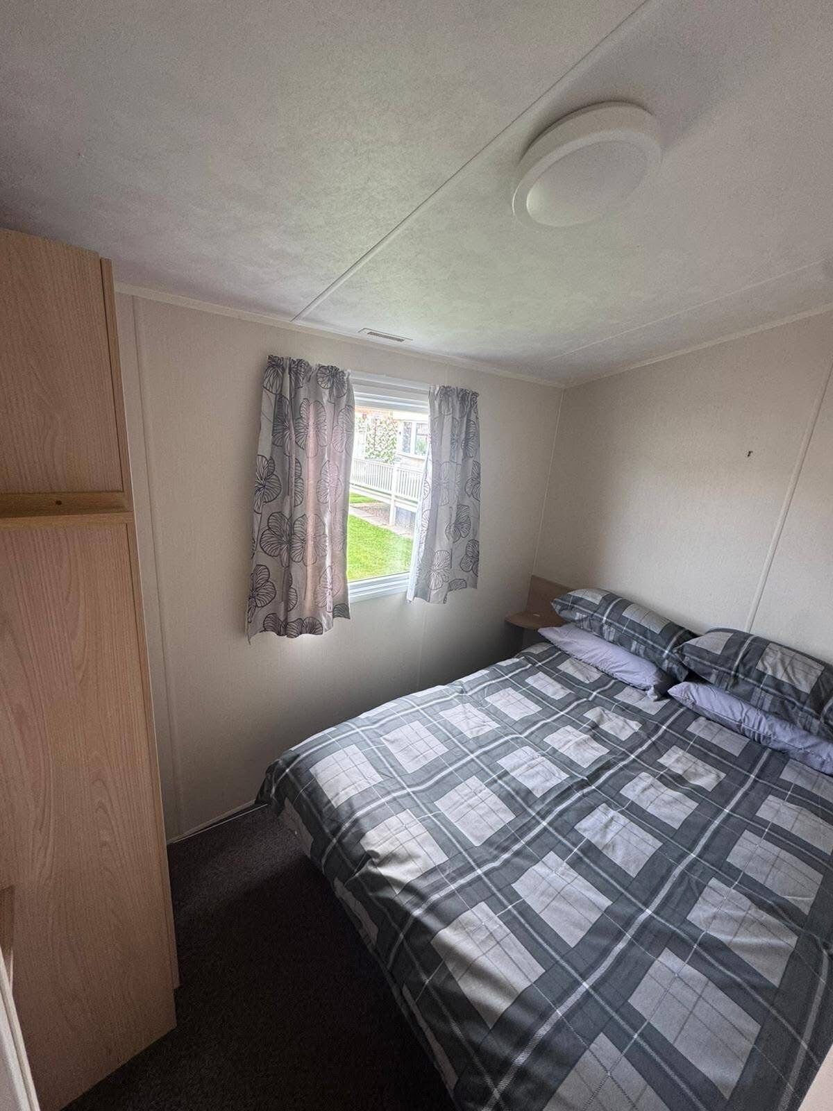 A small, single-bed room with a window, a wooden wardrobe, and a ceiling light fixture.