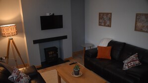 Ein Wohnzimmer mit schwarzem Ledersofa, einem Holztisch, einem Kamin, einem an der Wand befestigten Fernseher und einer Lampe.