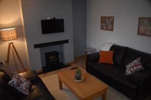 A living room with a black leather sofa, a wooden coffee table, a fireplace, a television mounted on the wall, and a lamp. - Snowdonia Riverside Cabin with Private River (Caernarfon)
