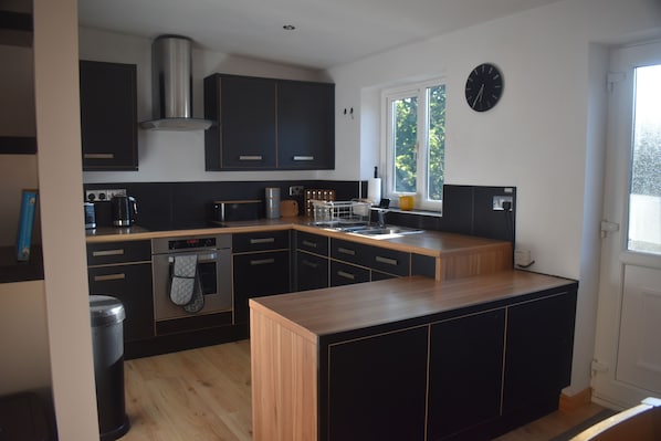 A modern kitchen with black cabinetry, a stainless steel range hood, a built-in oven, and a window with a view of greenery.