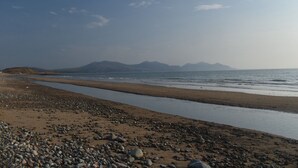 Ein Strand mit felsigem Ufer, ruhigem Wasser und Bergen im Hintergrund.
