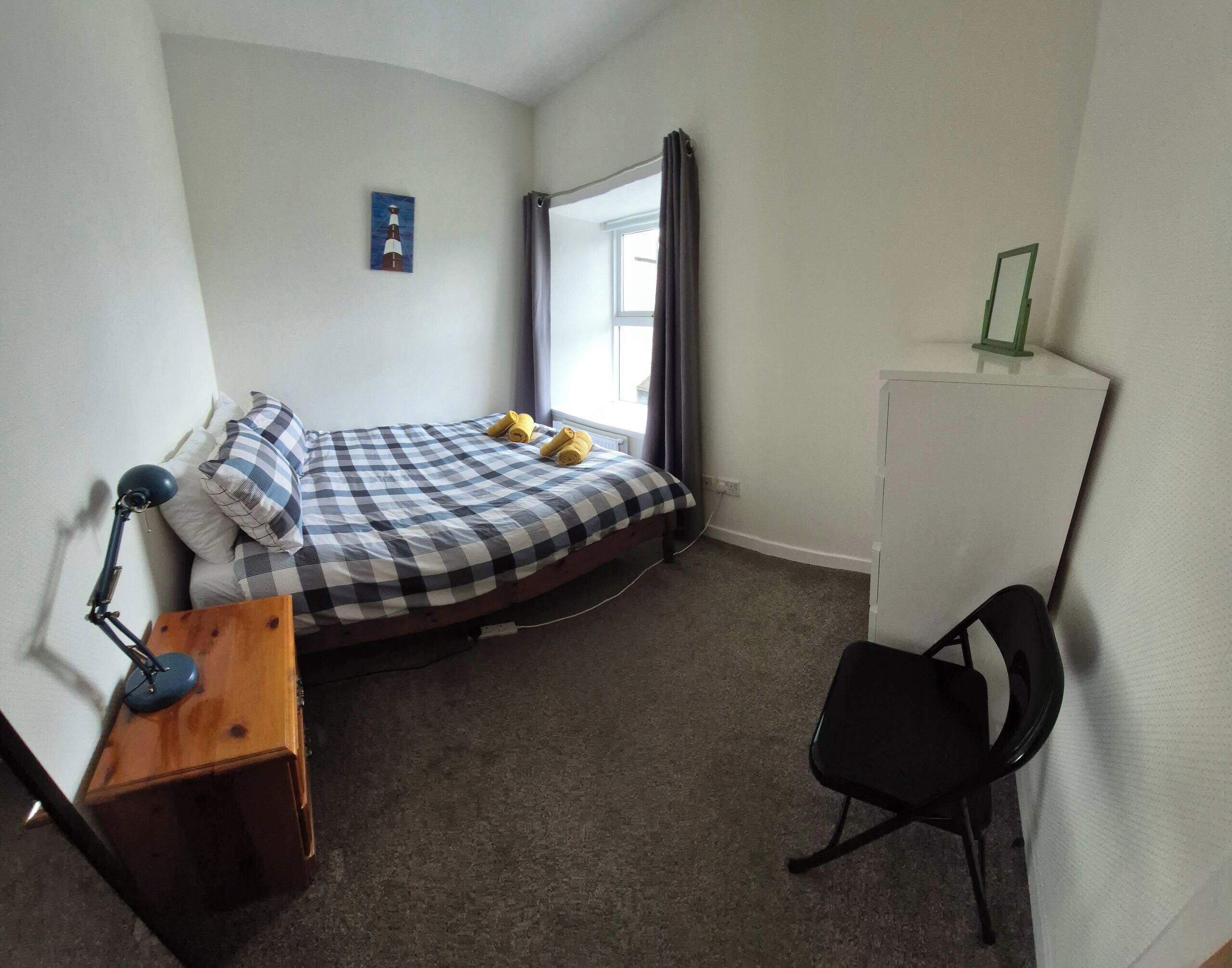 A small, single-bed room with a checkered bedspread, a desk with a lamp, a chair, and a small cabinet.