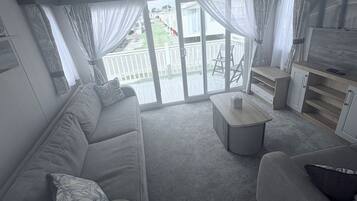 A modern living room with a grey sofa, a coffee table, and a TV. There is a sliding glass door leading to an outdoor deck.