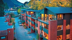 A modern multi-story residential building with balconies, a paved driveway, and a mountain backdrop.