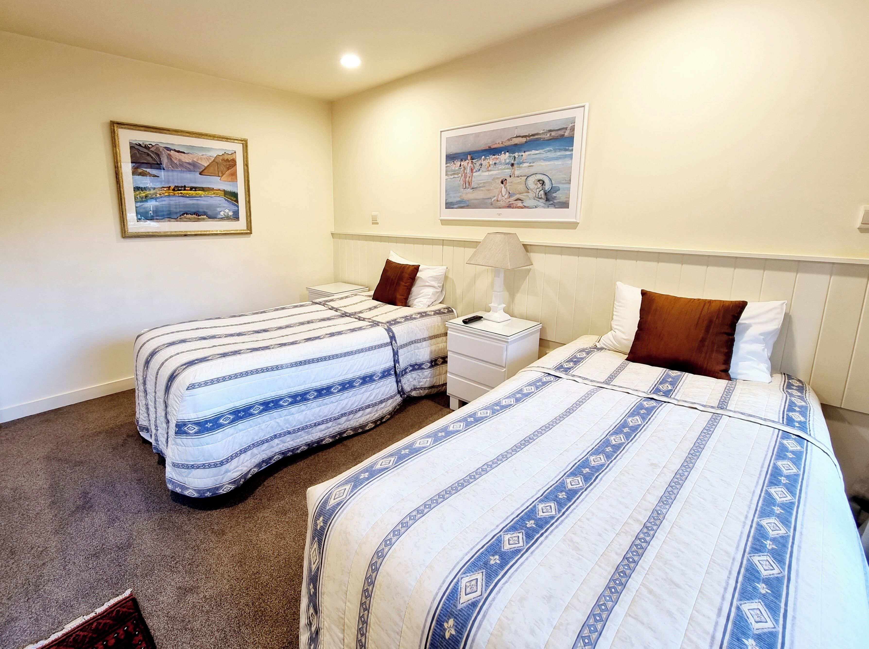 Habitación de hotel con dos camas, una mesita de noche con lámpara y dos cuadros en la pared.