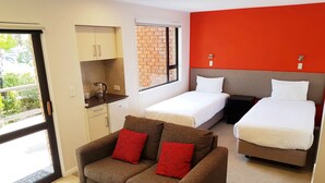 A room with a kitchenette, two beds, a sofa with red cushions, and a wooden table.