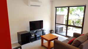 Una sala de estar con una televisión de pantalla plana, una mesa de centro de madera, un sofá y un ventanal corredero que da a un área exterior.