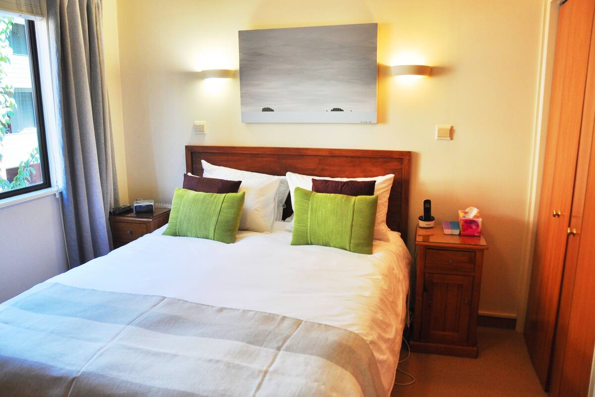 Una habitación de hotel con una cama grande, dos mesitas de noche, una lámpara y un cuadro en la pared, además de un ventanal con cortinas.