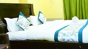 Una cama bien hecha con ropa blanca y detalles azules, un cabecero oscuro y una cortina verde al fondo.