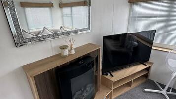 A compact living space with a television, a small refrigerator, a mirror, and a window with blinds.