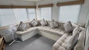 Ein kompakter Wohnbereich mit einer Sofagarnitur, einem Standventilator und einem Fenster mit Jalousien.