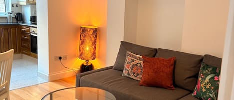 A modern living room with a sofa, a coffee table, and a lamp. There is a painting on the wall and a kitchen in the background.