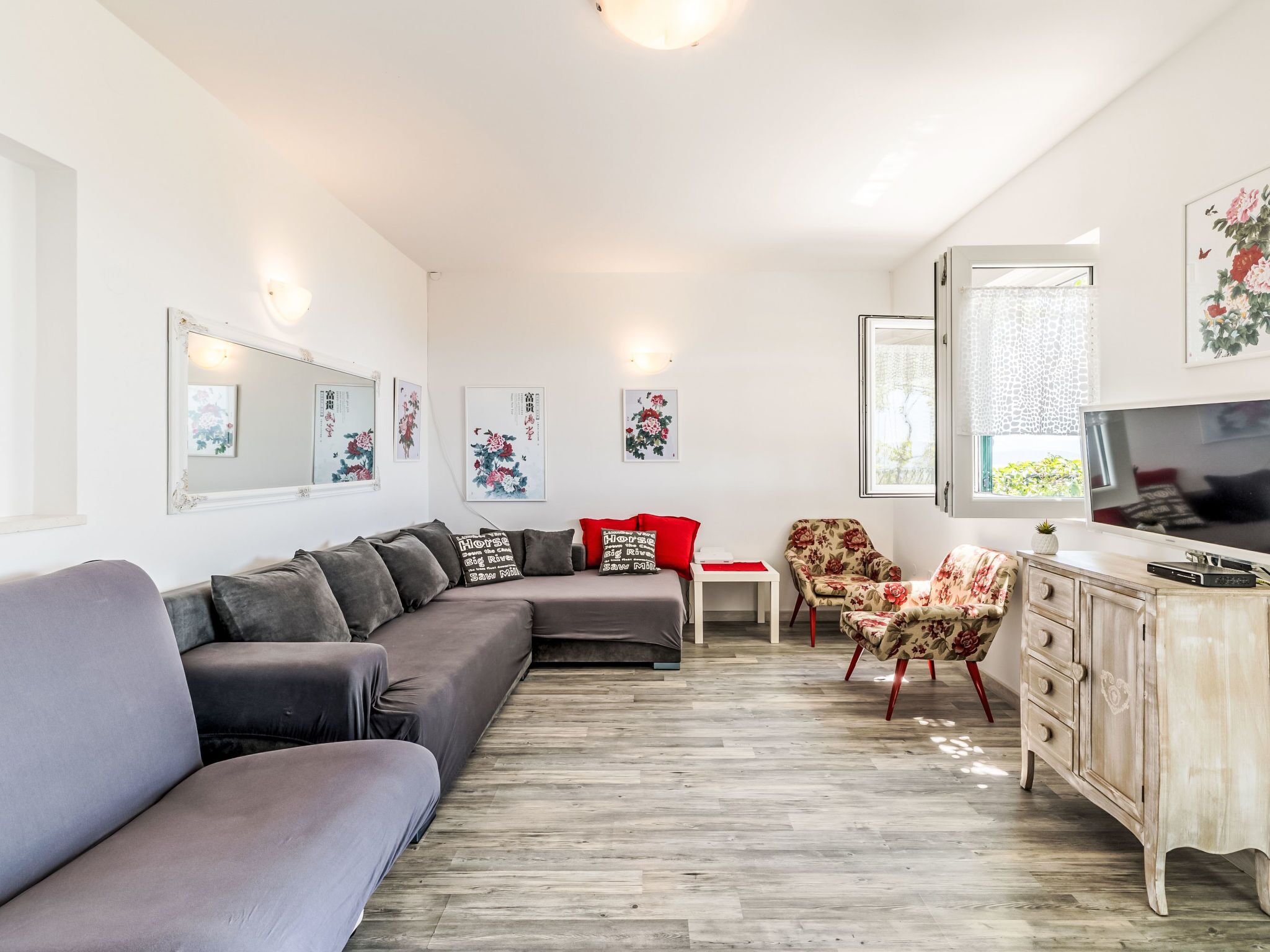 Ein modernes Wohnzimmer mit einem grauen Ecksofa, einem roten Sessel, einer Holzkommode und einem Fernseher.