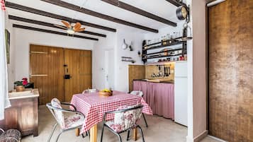 A dining area with a wooden table and chairs, a ceiling fan, and a kitchen area with shelves and utensils.