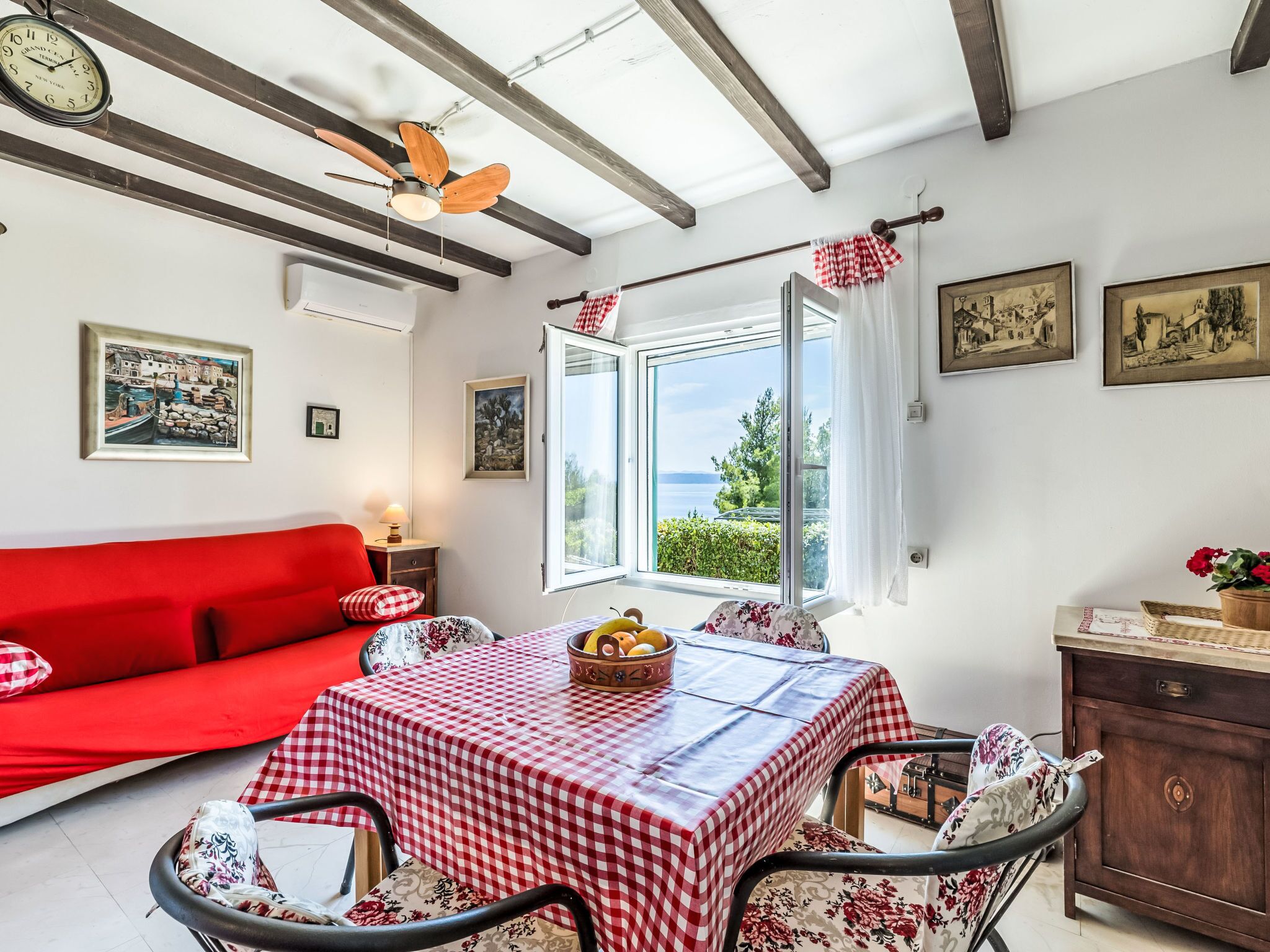 A room with a red sofa, a table with a checkered tablecloth, a ceiling fan, and a view through an open window.