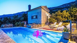 Una piscina con solárium de piedra, un edificio de piedra y montañas al fondo.