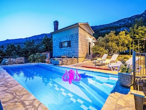 Una piscina con solárium de piedra, un edificio de piedra y montañas al fondo.