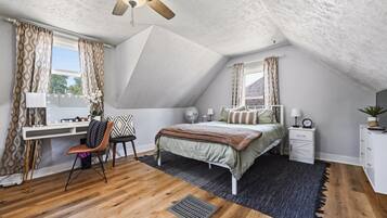 Un dormitorio con techo inclinado, una cama con cubrecama verde, un escritorio con silla y una mesita de noche con lámpara.