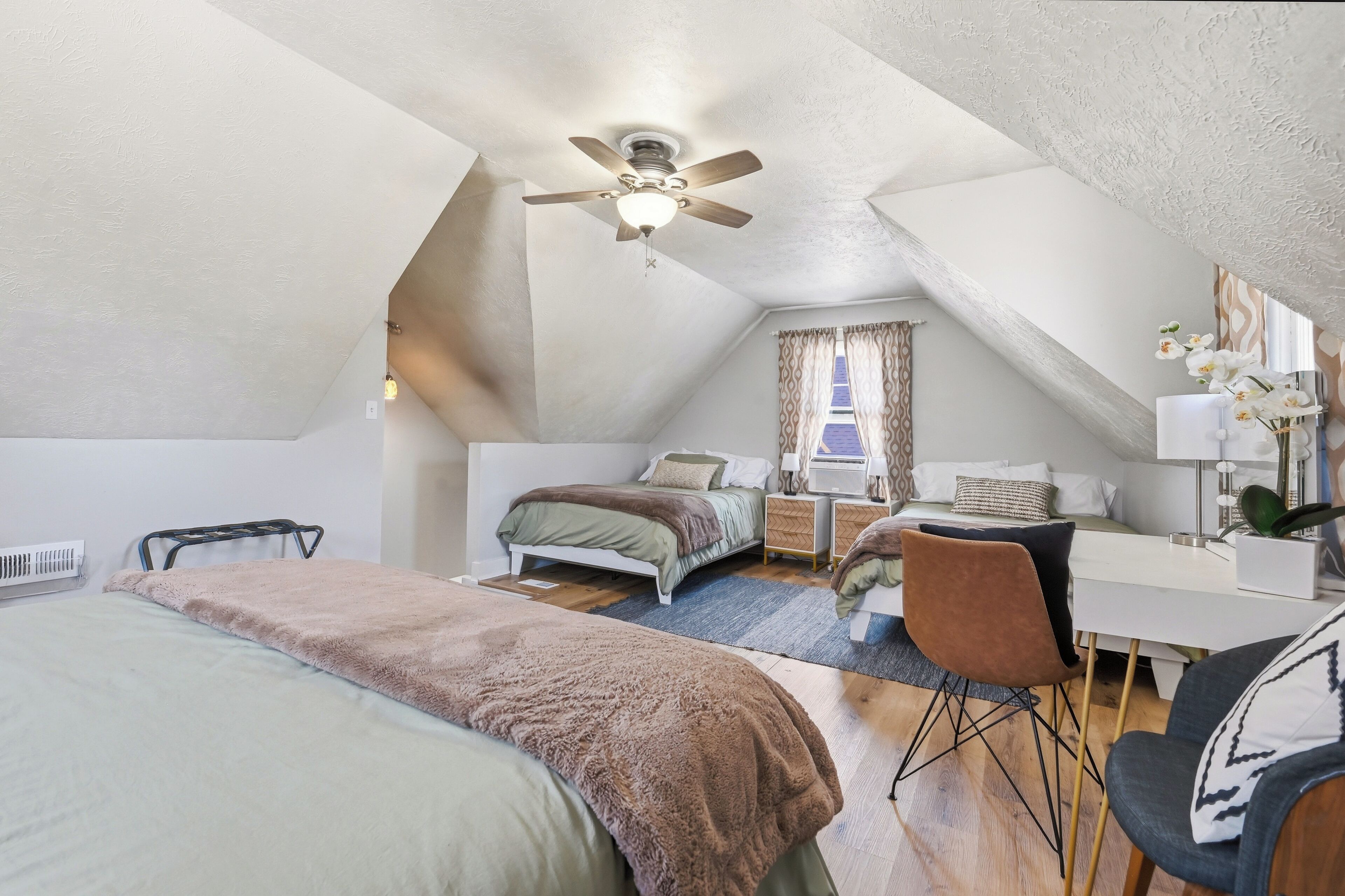 A bedroom with a bed, a desk, a chair, a ceiling fan, and a window with curtains.