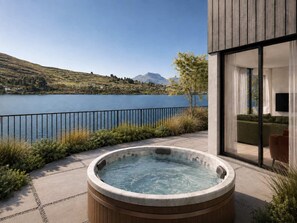 Un jacuzzi en un patio con vista a un lago, una terraza de madera y un edificio moderno con puerta de vidrio.