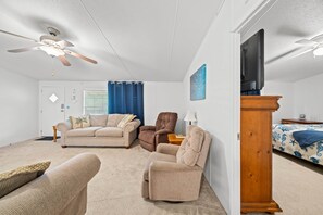 Een woonkamer met een plafondventilator, een televisie, een bank, een stoel en een bed met een decoratieve sprei.