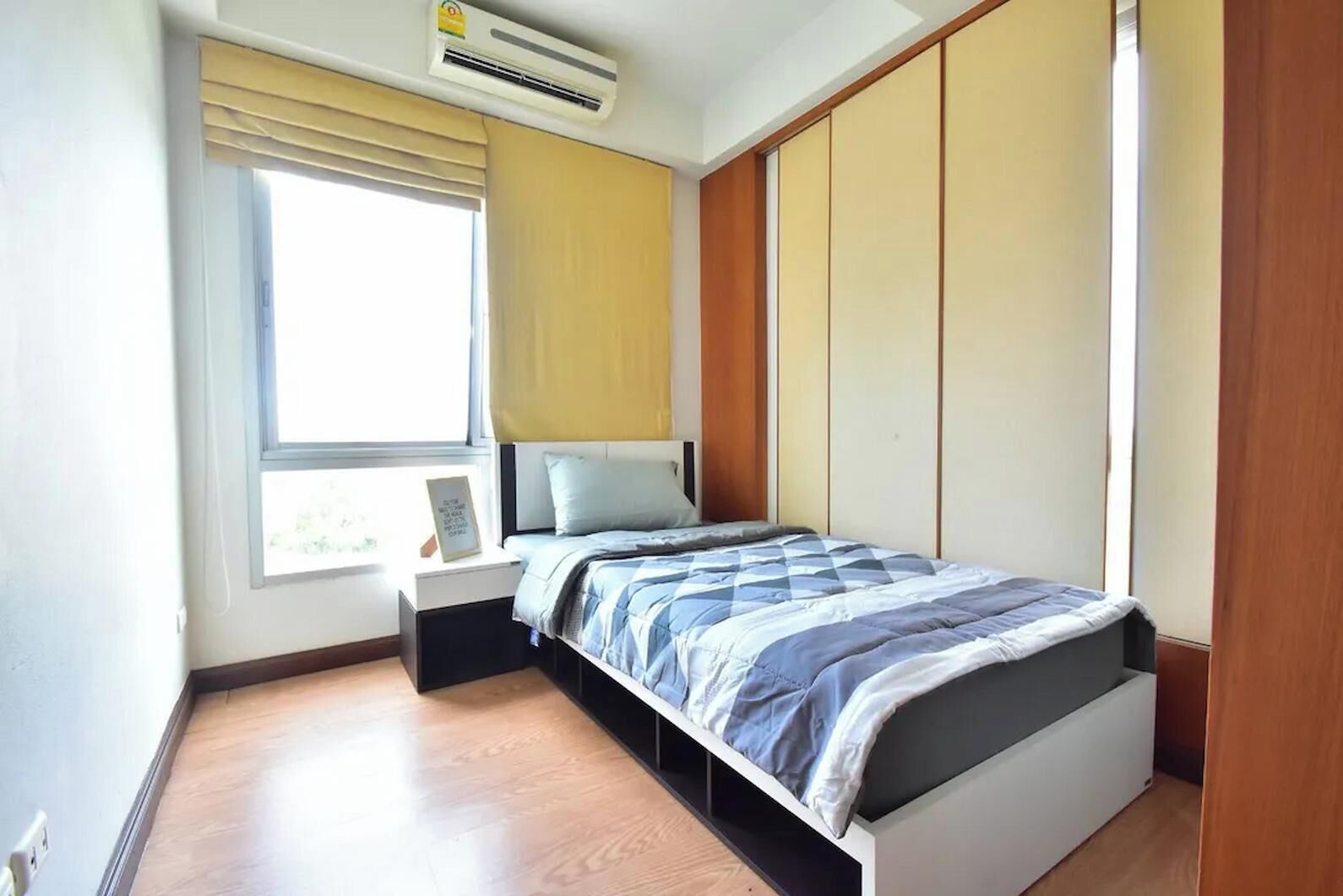 Un dormitorio con una cama, una mesita de noche, una ventana con cortinas y un equipo de aire acondicionado.
