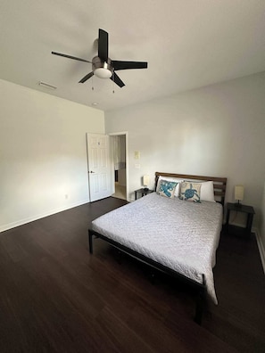 Un dormitorio con cama, ventilador de techo, una puerta y una mesita de noche con lámpara.