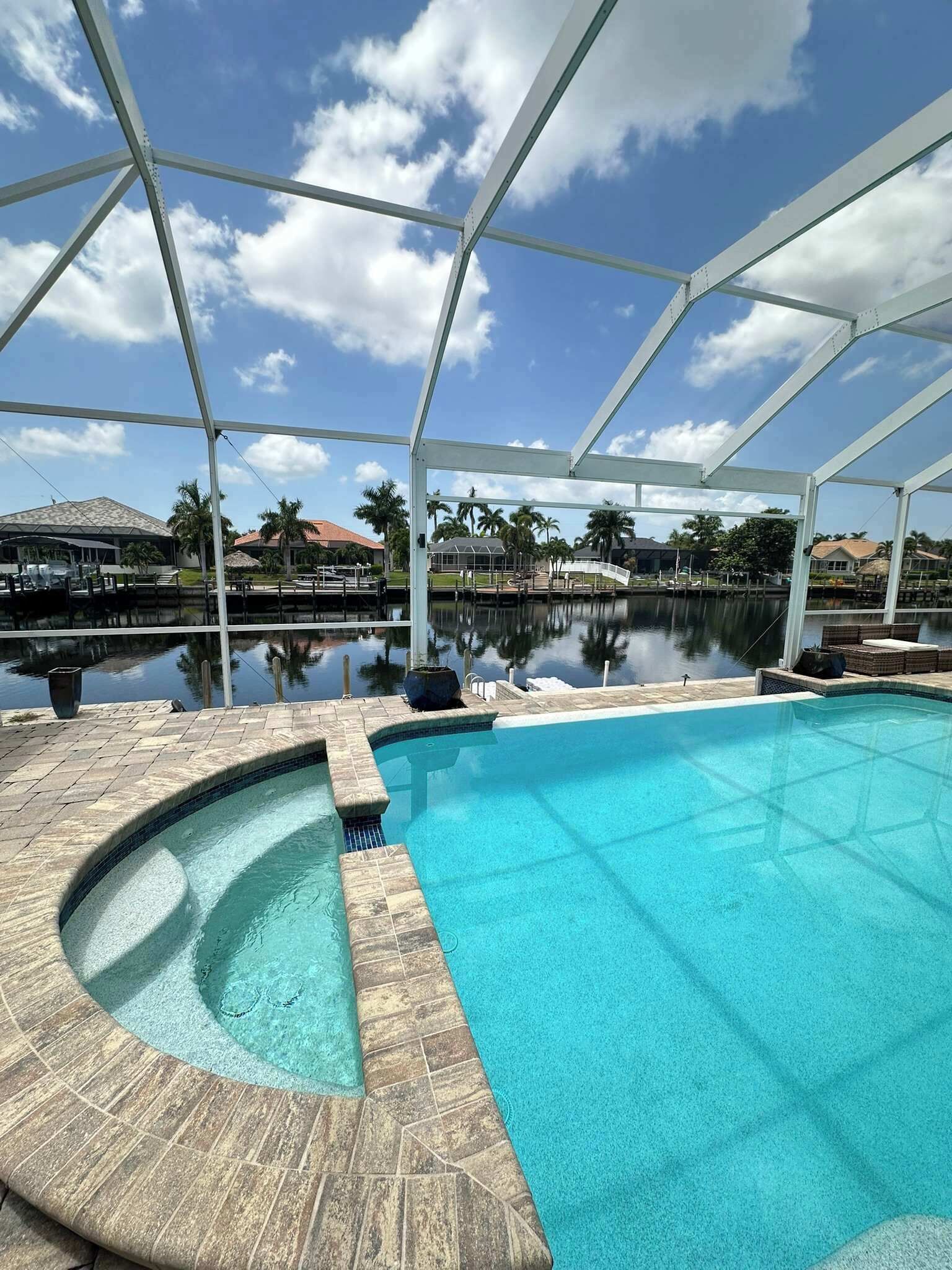 A swimming pool with a clear blue water feature, surrounded by a covered area with a view of a calm body of water and palm trees in the background.