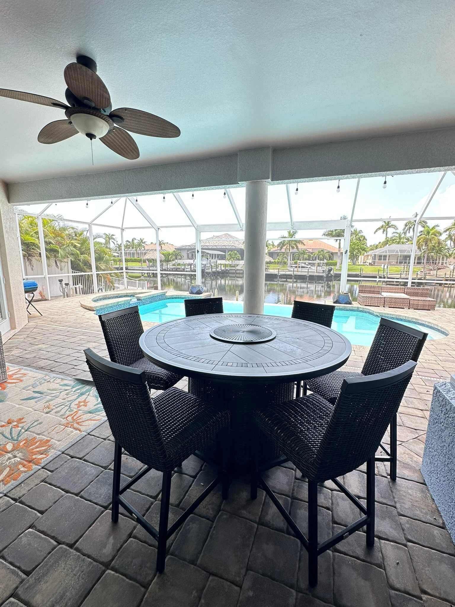 An outdoor patio with a round table and chairs, a ceiling fan, and a view of a swimming pool and buildings in the background.