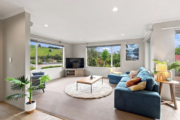 A spacious living room with a large TV, comfortable seating, and large windows offering a view of the outdoors.