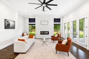 A modern living room with a fireplace, a ceiling fan, and a large window with blinds. - Houston’s Private Estate Venue | Weddings & Events (Houston)