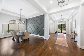 A spacious living area with a dining table and chairs, a large window with blinds, a geometric wall design, and a hallway leading to a door. - Houston’s Private Estate Venue | Weddings & Events (Houston)