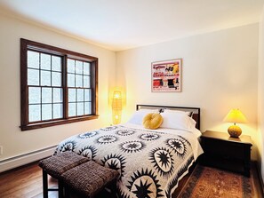 Quarto com uma janela grande, cama com colcha estampada, mesa de cabeceira com abajur e um pôster na parede.
