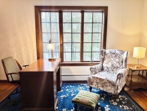 A room with a wooden desk, a chair, a patterned armchair, a small table with a lamp, and a view through a large window. - Private 7-Acre Retreat w/Brook, Near Hike & Ski (Townshend)