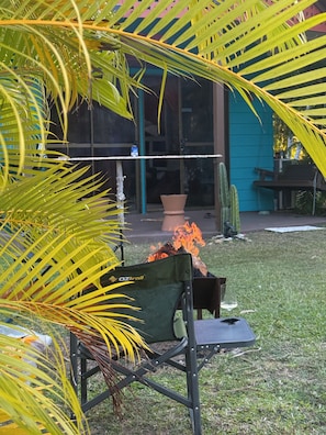 Un patio con una fogata, una silla plegable verde y un edificio de fachada azul al fondo.