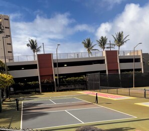 An outdoor tennis court with a net, surrounded by a fence, under a multi-level parking structure with palm trees. - Ocean View of Molokai  - 1-bedr / 1 bth remodeled on 6th floor Kaanapali Shores (Lahaina)