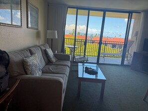 A living room with a sofa, a glass coffee table, a lamp, and a view of a balcony with a railing and greenery. - Ocean View of Molokai  - 1-bedr / 1 bth remodeled on 6th floor Kaanapali Shores (Lahaina)