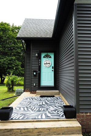 A house with a dark exterior, a light blue door with a welcome sign, and a patterned rug on the front porch.