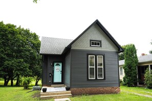 A small, single-story house with a dark siding exterior, a light blue door, and a small front porch.