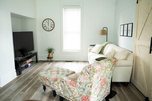 A living room with a sofa, a television, a clock, and a window with blinds.