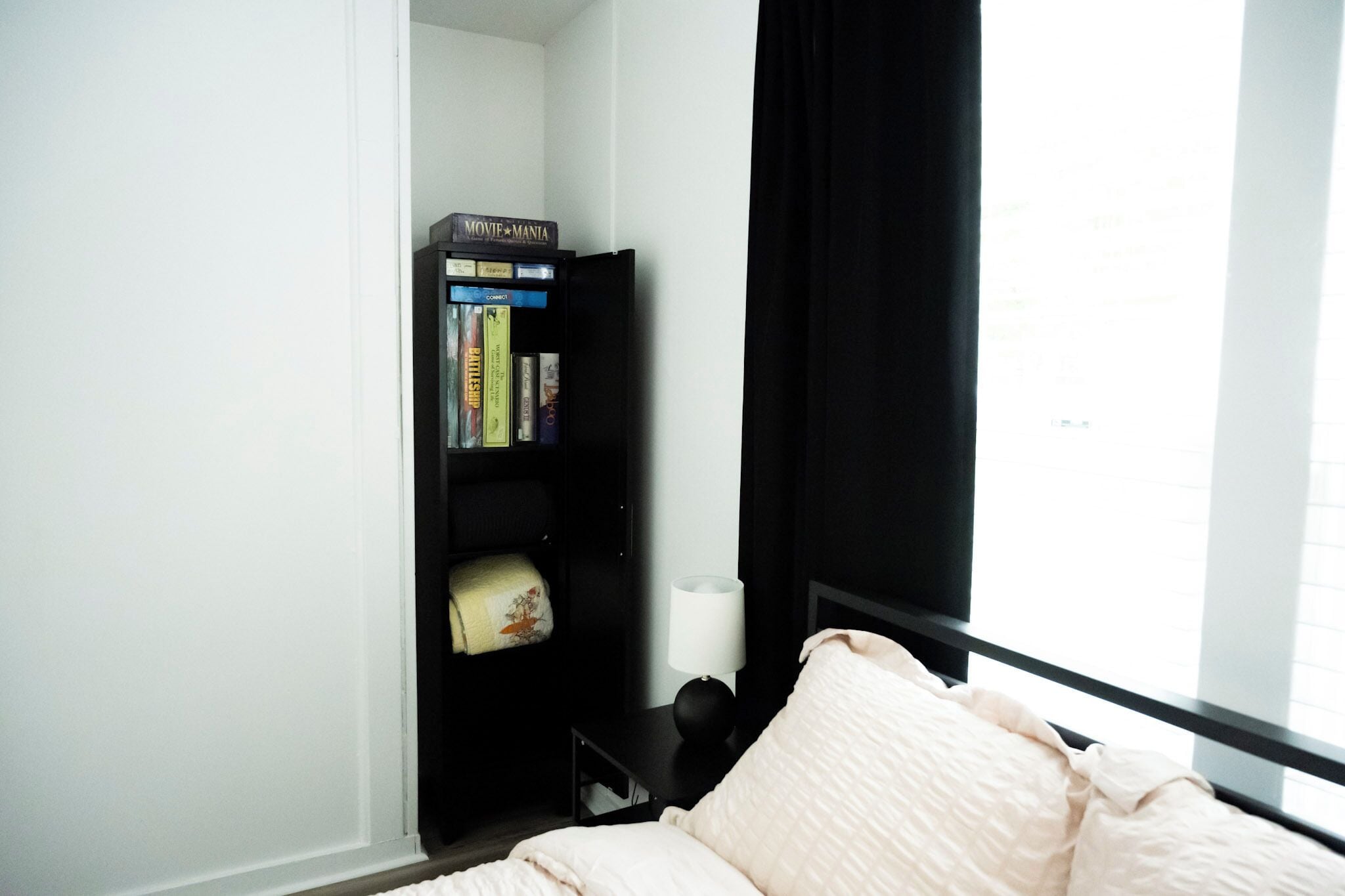 A bedroom with a bed, a bookshelf, a window with curtains, and a bedside table with a lamp.