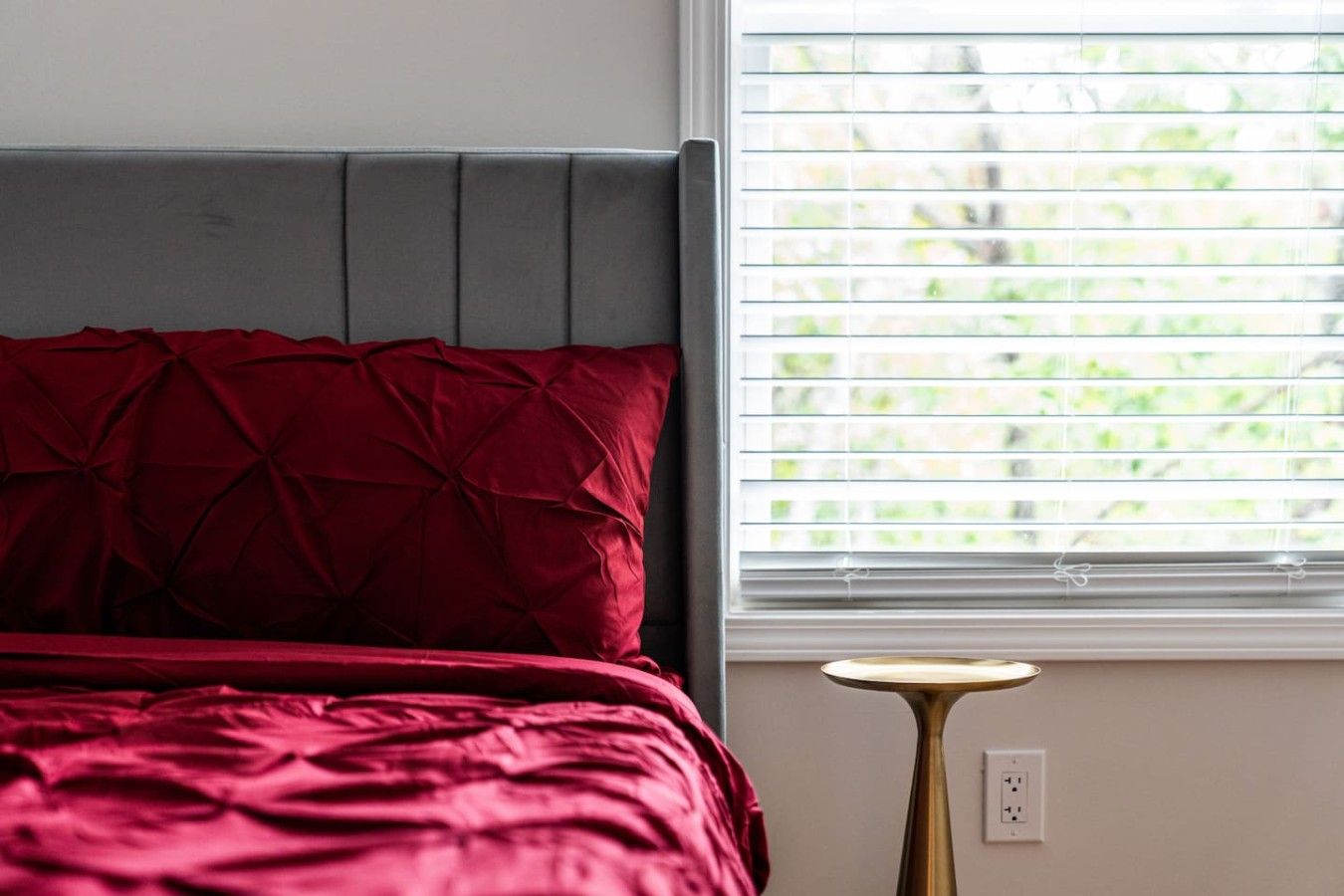 A bed with a grey headboard and red bedding, a bedside lamp, and a window with blinds.