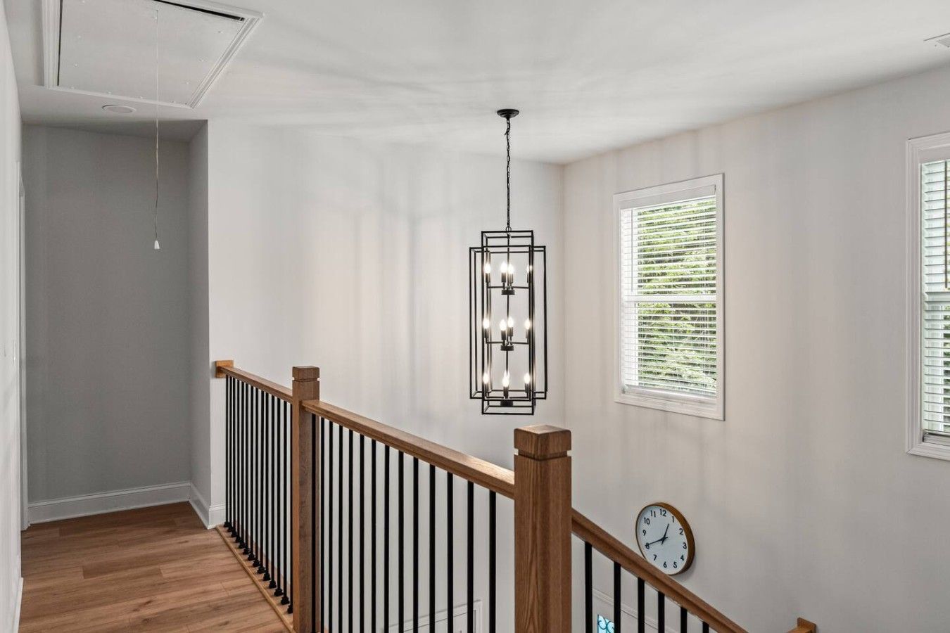 Un escalier avec une rampe en bois et des balustres verticales noires, menant à un palier où sont fixés un horloge murale et un luminaire suspendu.